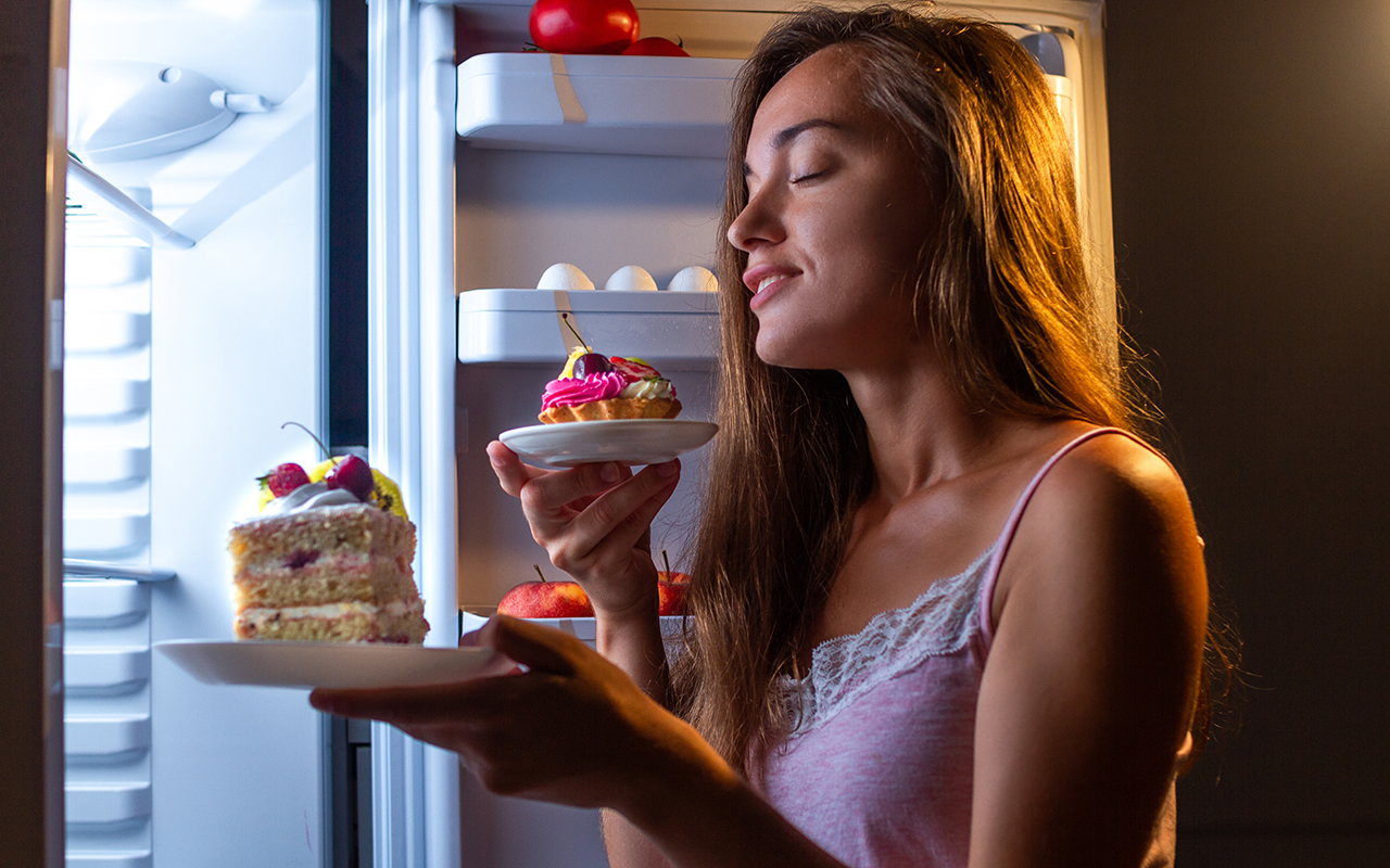 Девушка ест пончик возле холодильника. Голодная женщина. Есть по ночам. Девушка в белье ест пончик возле холодильника.
